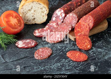 Würstchenstücke in Kreisen in der Nähe von Brot und Tomaten auf schwarzem Hintergrund geschnitten. Drei verschiedene Würste auf einem Schneidebrett. Weichfokus. Stockfoto