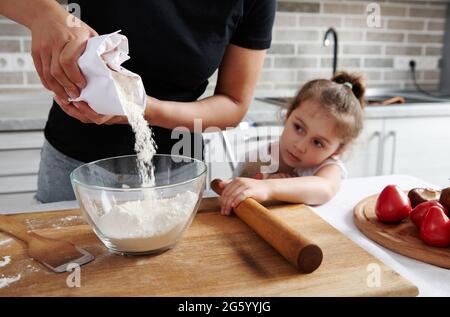 Eine Frau goss weißes Mehl aus Papierverpackungen in eine transparente Glasschüssel. Neben ihr sitzt ihre kleine Tochter und beobachtet den Kochvorgang. Stockfoto