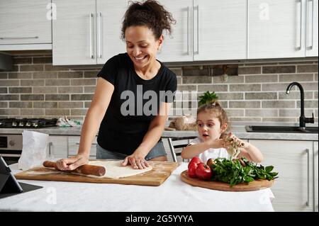 Mutter und Tochter kochen gemeinsam Pizza, Rollen den Teig mit einem Holzpfropfen aus und haben gemeinsam Spaß in der Küche Stockfoto