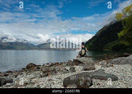 Ein Mann, der auf den Felsen sitzt und den Blick auf den Wakatipu-See in Queenstown genießt Stockfoto