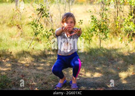 Defocus caucasian preteen Mädchen tun körperliche Bewegung im Park, Wald, im Freien, draußen. Gesunder Wellness-Lebensstil. Kniebeugen mit ausgestreckten Armen. Natur Stockfoto