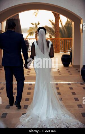 Rückansicht Aufnahme der schönen Braut und Bräutigam in wunderschönen Hochzeitskleid und schwarzen Anzug zu Fuß unter dem Bogen des Gebäudes Hände haltend Stockfoto