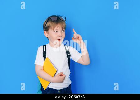 Zurück zum Schulkonzept. Schuljunge mit Rucksack und gelbem Buch vor blauem Hintergrund. Kind zeigt Daumen nach oben. Stockfoto