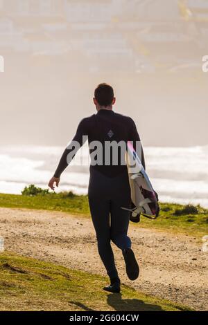 Die Rückansicht eines männlichen Surfers, der sein Surfbrett trägt und den Küstenpfad entlang in Richtung Fistral in Newquay in Cornwall läuft. Stockfoto