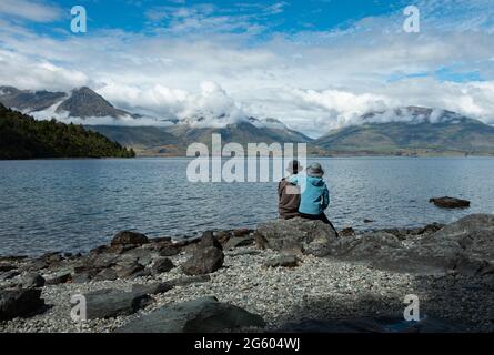 Ein Paar sitzt auf den Felsen und genießt den Blick auf die Berge über den Lake Wakatipu, Queenstown. Stockfoto