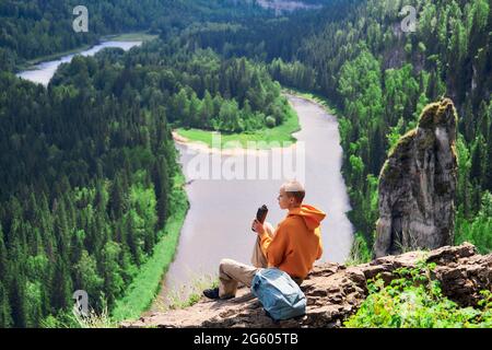 Teenager-Wandermädchen, die sich am Rand einer Klippe ausruhen und den Fluss vor dem Hintergrund einer natürlichen Landschaft betrachten Stockfoto