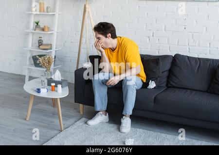 Kranker junger Mann, der zu Hause auf der Couch sitzt und mit der Hand in Gesichtsnähe hustet Stockfoto