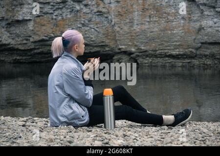 Teenager-Wandererin, die sich mit einem Becher am Flussufer ausruhte und auf die Küstenfelsen schaute Stockfoto