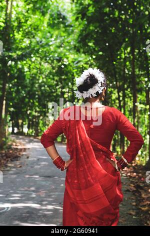 Junge Frau mit rotem Saree, die auf einer grünen Asphaltstraße unterwegs ist. Das Mädchen geht den Weg durch den Wald bis zum Licht. Stockfoto