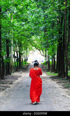 Junge Frau mit rotem Saree, die auf einer grünen Asphaltstraße unterwegs ist. Das Mädchen geht den Weg durch den Wald bis zum Licht. Stockfoto