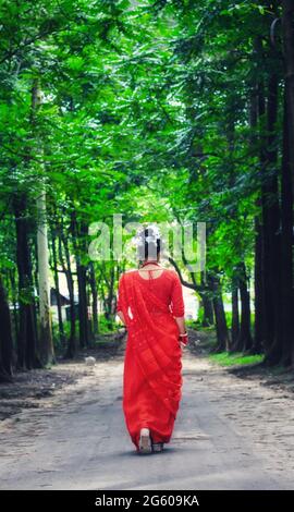 Junge Frau mit rotem Saree, die auf einer grünen Asphaltstraße unterwegs ist. Das Mädchen geht den Weg durch den Wald bis zum Licht. Stockfoto