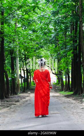 Junge Frau mit rotem Saree, die auf einer grünen Asphaltstraße unterwegs ist. Das Mädchen geht den Weg durch den Wald bis zum Licht. Stockfoto