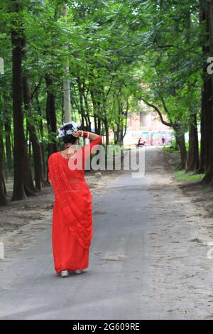 Junge Frau mit rotem Saree, die auf einer grünen Asphaltstraße unterwegs ist. Das Mädchen geht den Weg durch den Wald bis zum Licht. Stockfoto