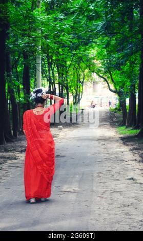 Junge Frau mit rotem Saree, die auf einer grünen Asphaltstraße unterwegs ist. Das Mädchen geht den Weg durch den Wald bis zum Licht. Stockfoto