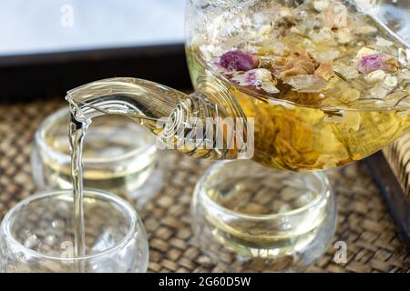 Kräutertee der trocknenden Blumen gießt in Tasse, Nahaufnahme. Eine Teekanne und Tasse mit Blumen Tee auf dem Tisch. Stockfoto