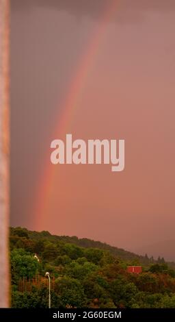 Regenbogen steigt aus dem Wald mit dunklen und dichten Regenwolken im Hintergrund - eingerahmt von verschwommenen Hauswänden Stockfoto