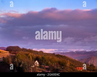 Hügelige Landschaft mit schneebedeckten Hügeln in der Ferne am Horizont und dichten Wolken am Himmel. Von der Sonne beleuchtete Landschaft vor Sonnenuntergang. Stockfoto