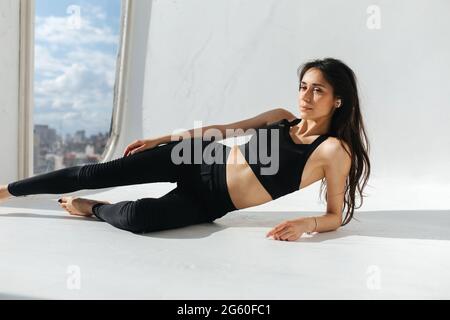 Schlanke armenische Frau in schwarzer Sportkleidung, meditiert auf weißem Boden in der Nähe des Fensters Stockfoto