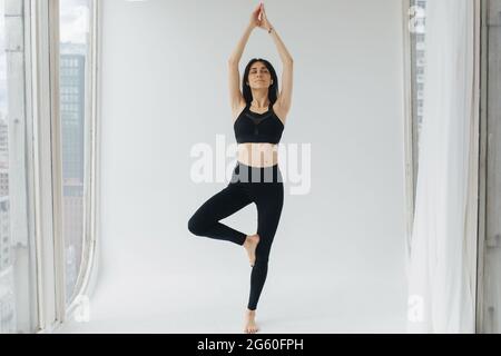 armenische Frau in schwarzer Sportkleidung, die mit erhobenen Händen die Baumhaltung übt Stockfoto