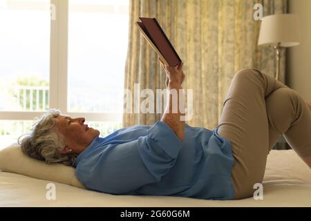 Ältere kaukasische Frau, die im Bett lag und im Schlafzimmer ein Buch las Stockfoto