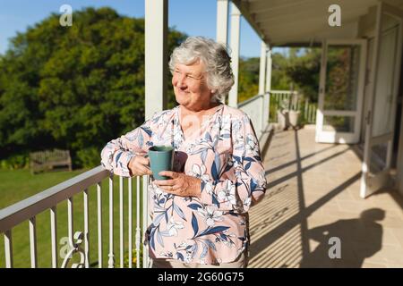 Ältere kaukasische Frau, die auf dem Balkon steht und den Becher hält und lächelt Stockfoto