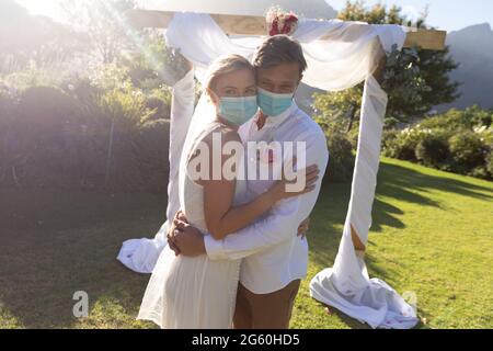 Glückliche kaukasische Braut und Bräutigam heiraten in Gesichtsmasken und umarmen Stockfoto