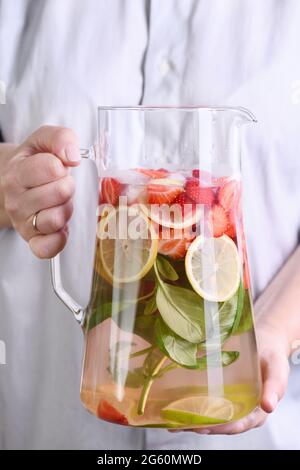 Eine Kanne mit einem erfrischenden Bio-Softdrink mit reifen frischen Erdbeeren, Zitronenscheiben mit Limette und Basilikum, in weiblicher Nahaufnahme. Stockfoto