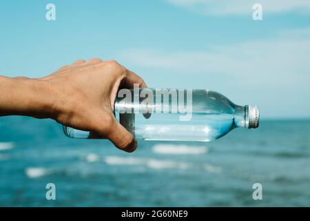 Nahaufnahme eines jungen kaukasischen Mannes, der eine wiederverwendbare Glasflasche mit Wasser in der Hand hält, vor dem Ozean, ausgerichtet am Horizont Stockfoto
