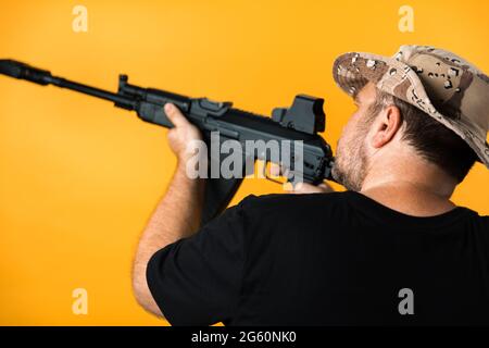 Mann mittleren Alters mit Gewehr in schwarzem T-Shirt und Hut vor gelbem Hintergrund. Stockfoto
