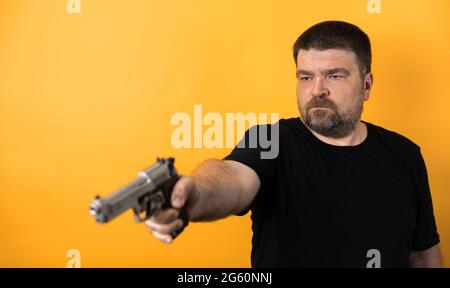 Mann mittleren Alters mit Pistole in schwarzem T-Shirt auf gelbem Hintergrund. Stockfoto