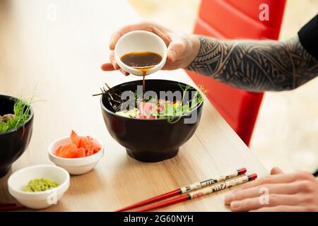 Ein Mann fügt dem Poke-Salat Sojasauce hinzu. Eine Männerhand gießt Sojasauce in eine Schüssel Salat. Junger Mann, der in einem Restaurant oder Café Salat mit Sauce zubereitet Stockfoto