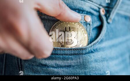 Nahaufnahme von Männerfingern, die die Bitcoin-Münze in die blaue Jeans-kleine Tasche stecken. Ersparnisse, digitale Währung, Kryptowährung, Investitionen und s Stockfoto