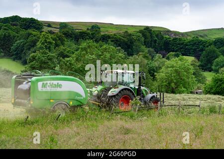 Heu- und Silageherstellung (Landwirt im Traktor auf dem Bauernhof bei der Arbeit auf landschaftlich reizvollen Feldern, sammelt trockenes Gras, wickelt Rundballen in Ballenpresse) - Yorkshire England, Großbritannien. Stockfoto
