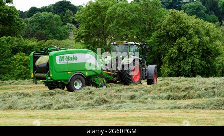 Heu- oder Silageherstellung (Landwirt im Traktor auf dem Bauernhof bei der Arbeit auf dem Land, beim Sammeln von trockenem Gras, beim Einwickeln von Rundballen in Ballenpressen) - Yorkshire England, Großbritannien. Stockfoto