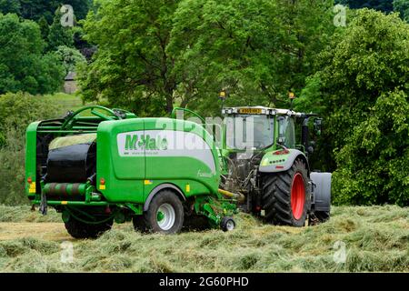 Heu- oder Silageherstellung (Landwirt im Traktor auf dem Bauernhof bei der Arbeit auf dem Land, beim Sammeln von trockenem Gras, beim Einwickeln von Rundballen in Ballenpressen) - Yorkshire England, Großbritannien. Stockfoto