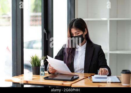 Geschäftsfrau trägt Maske, während sie im Büro Taschenrechner mit Computer-Laptop, Geschäftsbuchhaltung, Budget- und Leihpapier verwendet Stockfoto