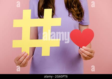 Nahaufnahmen von zugeschnittenen Aufnahmen einer jungen Frau, die ein T-Shirt trägt, zeigen ein großes gelbes Hashtag-Schild und ein kleines rotes Herz, das isoliert über einer rosa Wand posiert Stockfoto