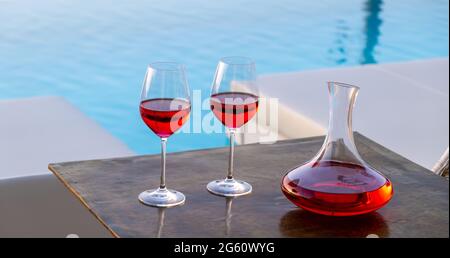 Rotweingläser am Swimmingpool. Zwei Weingläser und Dekanter auf Holztisch, türkisblauer Wasserhintergrund. Entspannung und Romantik durch die RE Stockfoto