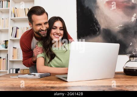 Lächelnder Mann umarmt Freundin mit Laptop zu Hause Stockfoto