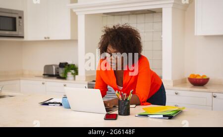 afroamerikanische Frau, die in der Küche arbeitet, sich am Laptop auf die Theke lehnt und sich konzentriert Stockfoto