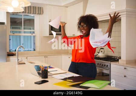 Glückliche afroamerikanische Frau, die mit einem Laptop in der Küche arbeitet, Erfolge feiert und Papiere wirft Stockfoto