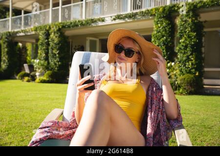 Lächelnde kaukasische Frau, die im sonnigen Garten mit Sonnenhut und Sonnenbrille auf dem Smartphone sitzt Stockfoto