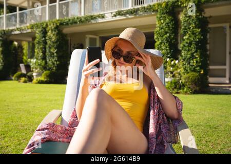 Kaukasische Frau, die im sonnigen Garten sitzt und Sonnenhut und Sonnenbrille mit dem Smartphone trägt Stockfoto