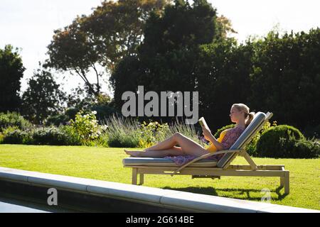 Lächelnde kaukasische Frau, die sich auf einer Sonnenliege im wunderschönen sonnigen Garten entspannt und ein Buch liest Stockfoto