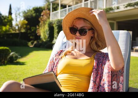 Kaukasische Frau mit Sonnenhut und Sonnenbrille, die sich im sonnigen Garten entspannt und ein Buch hält Stockfoto