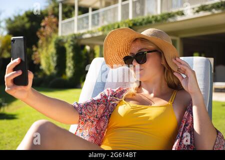 Kaukasische Frau, die im sonnigen Garten sitzt, trägt Sonnenhut und Sonnenbrille und nimmt Selfie mit dem Smartphone auf Stockfoto