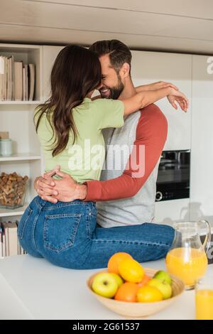 Lächelnder Mann umarmte Freundin neben Orangensaft und verschwommenes Obst in der Küche Stockfoto