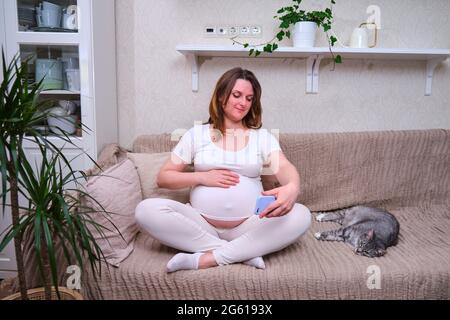 Videos im Internet aufnehmen, wenn eine Frau auf einem Heimbett schwanger ist. Moderne Technologien im Telefon bei Schwangeren Stockfoto