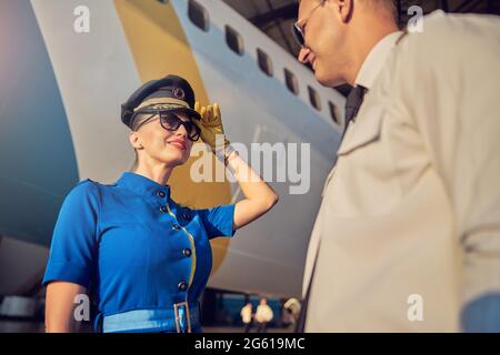 Charmante Frau Flugbegleiterin in Hut und Anzug begrüßen Pilot in der Nähe des Flugzeugs Stockfoto