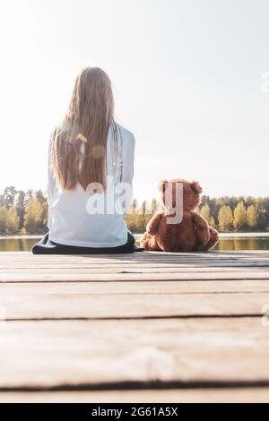 Kleines Mädchen mit Teddybär sitzt am Pier in der Nähe des ruhigen Flusses Stockfoto